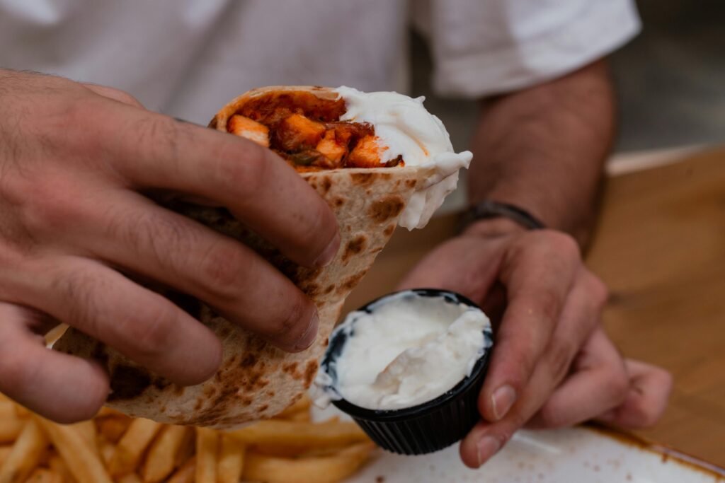 A close-up of a person dipping a shawarma wrap in sauce, with fries on the side.